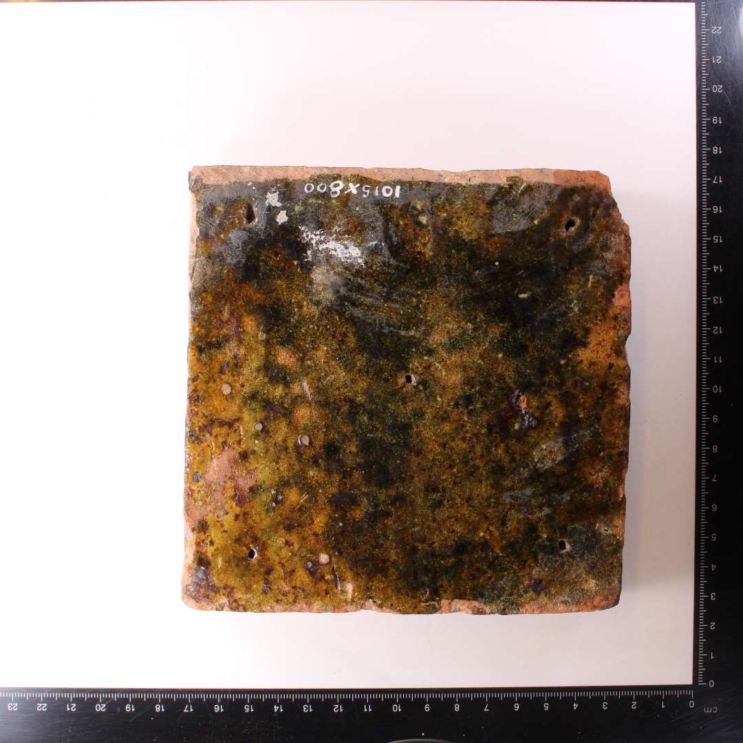 Kvadratisk gulvflise. Mørkegrøn glasur. 13.5x13,5 cm. tykkelse: 2,8 cm. Pindestik i hjørner og midt på. Samt to fragmentr af samme type og tykkelse