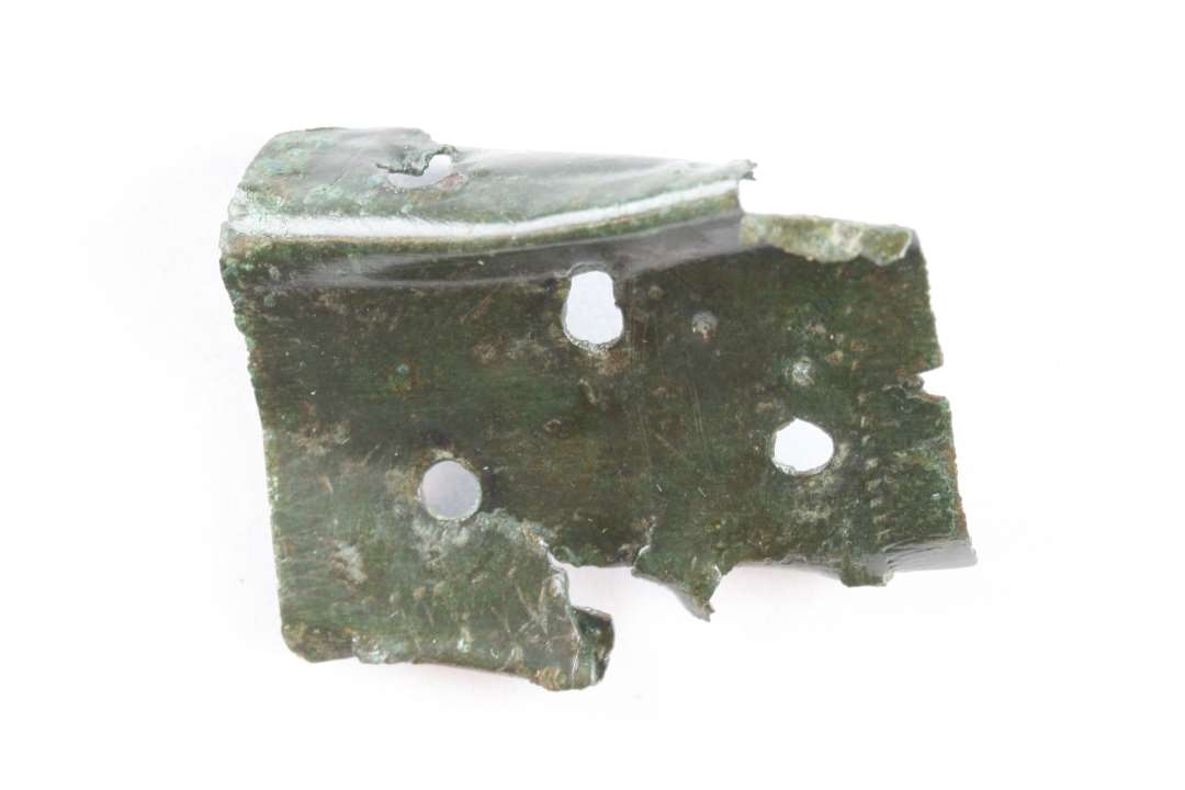 Fragmenteret beslag af kobberlegering. Med to ombuk og mindst re nittehuller. Dekoreret med indpunslet zig zagmønster. Mål: 3,6x2,5 cm.