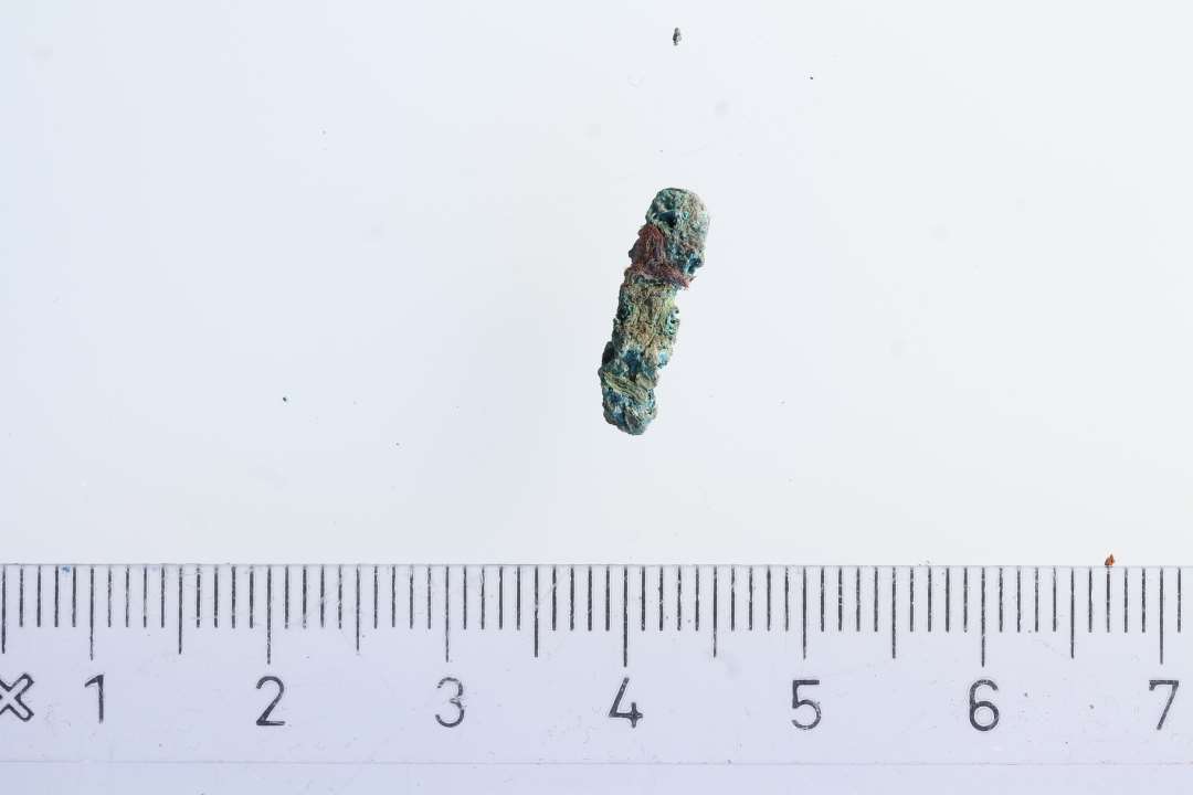 Lille stykke bronze på ca. 1,2 cm. En mulig torn fra et dragtspænde, med et meget lille stykke rød/lilla tekstil, der synes at være på begge sider.  