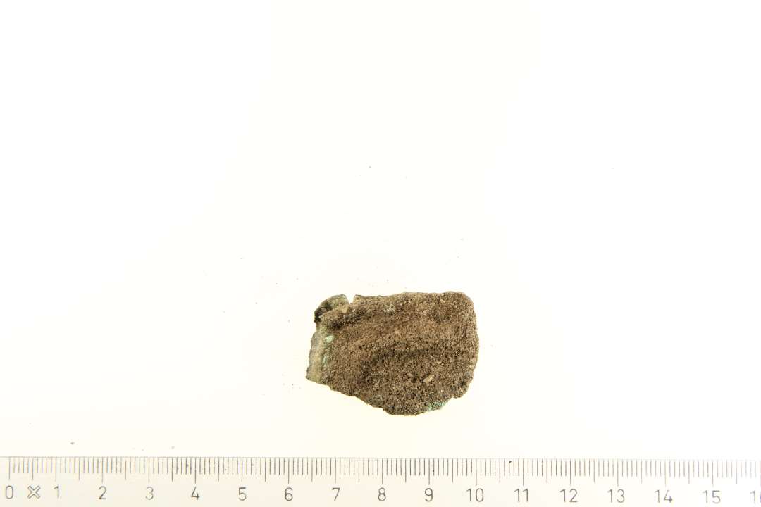 Bronze genstand på ca. 2-2,5 cm. Tolkes til at være et beslag. På den øverste side af genstanden, ses 3 fordybninger af forskellig størrelse. Udfra hvad der minder om en slags rand på undersiden af genstanden, kan det muligvis også være en beholder.