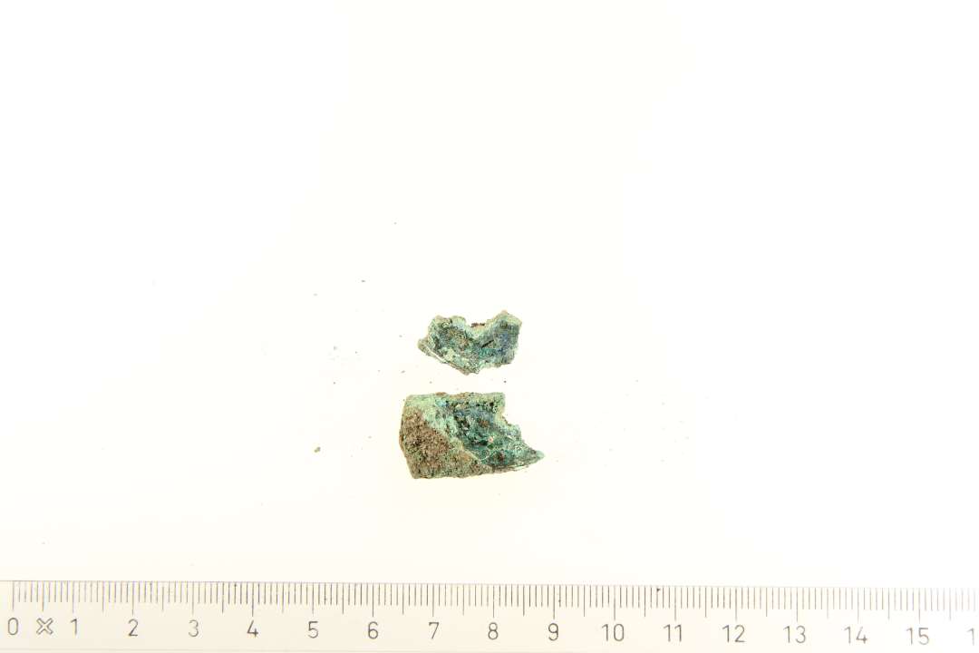 To små genstande af bronze, formentlig fragmenter af samme genstand. Den største af gentandene er lukket sammen i den ene side og nedbrudt i den anden. Der ses fine nittermærkninger, og kan tolkes som et fragment af et remendebeslag.