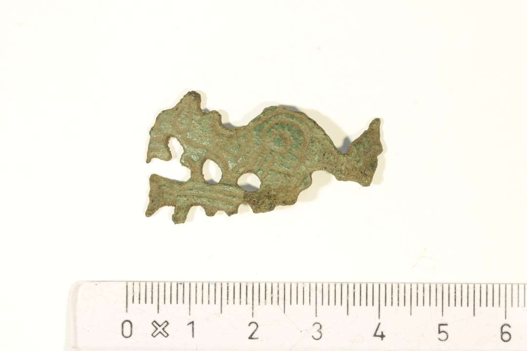 Fragment af dyreformet fibel af bronze ornamenteret med geometrisk stregornamentik, måske urnesstil? Dyret har mandelformet øje og spiralornament på skulderen. Dyret er højrestillet og ses i profil. Der er svage rester af nåleanordning på bagsiden. Mål: 4x2,1 cm.