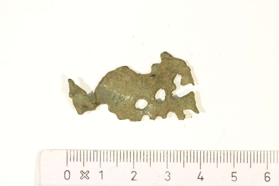 Fragment af dyreformet fibel af bronze ornamenteret med geometrisk stregornamentik, måske urnesstil? Dyret har mandelformet øje og spiralornament på skulderen. Dyret er højrestillet og ses i profil. Der er svage rester af nåleanordning på bagsiden. Mål: 4x2,1 cm.
