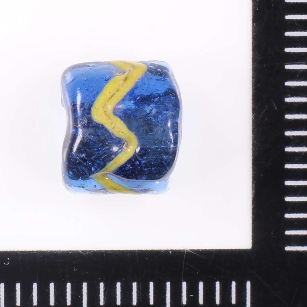 Halv cylindrisk gennemsigtig blå lidt kantet perle med gul tråd lagt i sigsag. Værktøjsspor ved hvert 