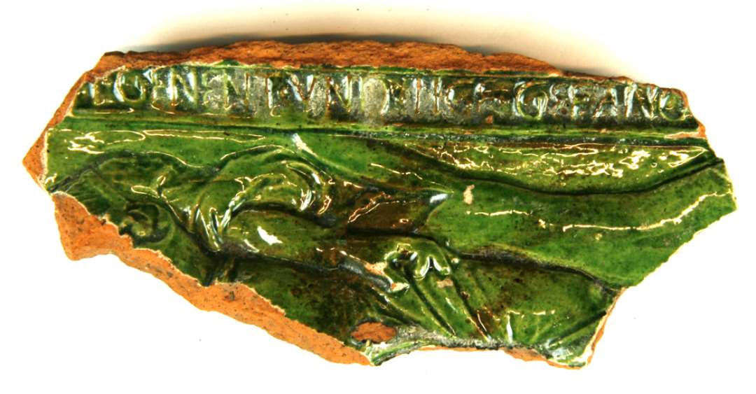 Fragment af kakkel af rødbrændt lergods med ydersiden dækket af pibelersbegitning, hvorover grøn blyglasur. Langs randen af den afbrudte yderramme findes et indskriftbånd med latinske bogstaver, hvoraf kan tydes: GENENT VND ?GE GEFANG ---- Af kaklens midterfelt er et smalt parti bevaret, der viser en del af et kvindelegeme hyllet i folderigt gevandt. Kvindens hånd synes lænket til en ræv eller hund ved hendes side. Største mål: 13 cm.