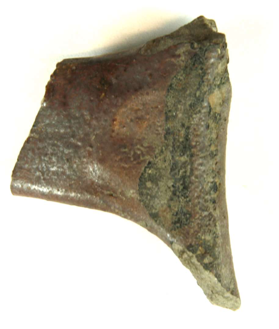 Hankebasisfragment af gråbrændt finslemmet stengods, hvis overflade er dækket af manganbrunlig bemaling, hvorover saltglasur. Hanken har bøjleform med fladt ovalt tværsnit.