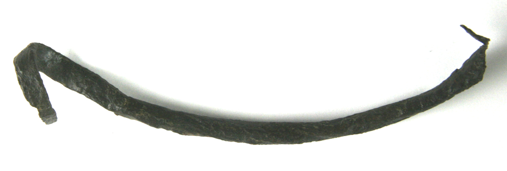 Fragment af hank af jern. Hanken har haft omtrent halvcirkulær form og er fremstillet af en trind jernstang, der er udformet med en grisehalekrølle i den ene ende, medens den anden ende, der har dannet hankens midterparti, er udhamret i flad form med rundet underside og hulkehl på oversiden. Største diagonale længdemål: 14,8 cm.