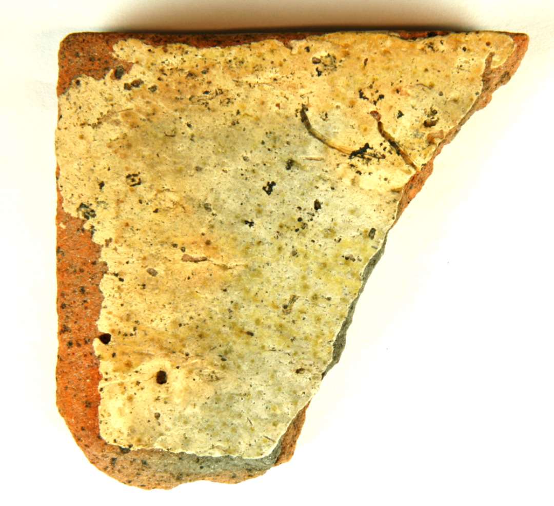 Hjørnefragment af gulvflise af rødbrændt lergods. Oversiden, der er stærkt afslidt, er dækket af et lag hvid pibelersmasse, hvorover anes rester af klar til grønlig blyglasur.  Største længde og bredde: ca. 7,2 cm. Tykkelse: 1,6 cm.