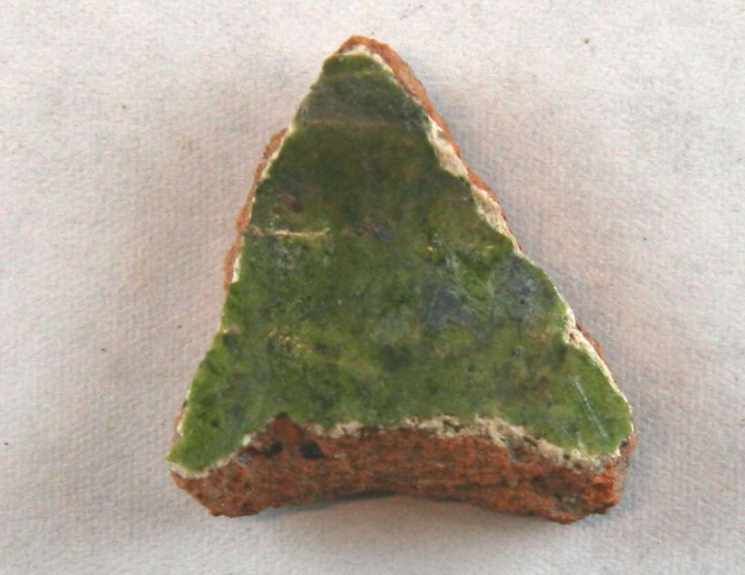 Halsskår af rødbrændt lergods med pibelersbegitning på ydersiden, der er dækket af grønlig blyglasur. På indersiden findes spredte pletter af klar blyglasur.