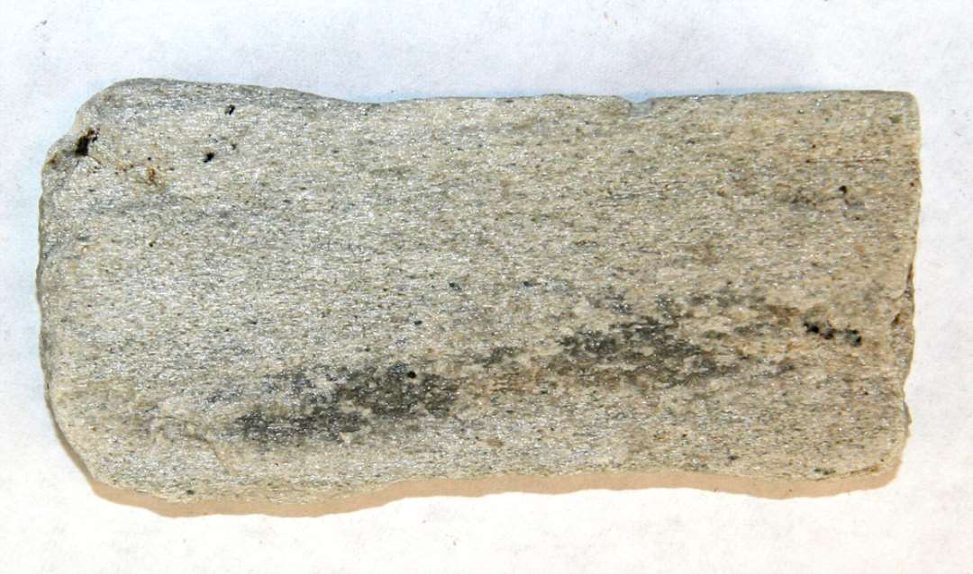 1 fladt aflangt stykke lysegråt hvæssestensfragment af sandsten afbrudt i begge ender. I den ene langsidekant ses halvdelen af en konisk ca. 2 mm dyb nedboring. Stenen synes iøvrigt uforarbejdet. Længde: ca. 8 cm. Bredde: 3,8 cm. Tykkelse: ca. 1 cm.