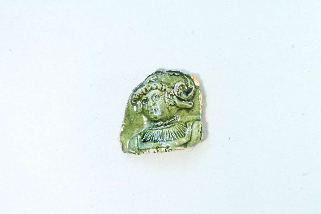 Fragment af grønglaseret kakkel. Der ses et hoved, der kunne være en kvinde. Største mål ca. 4 cm.