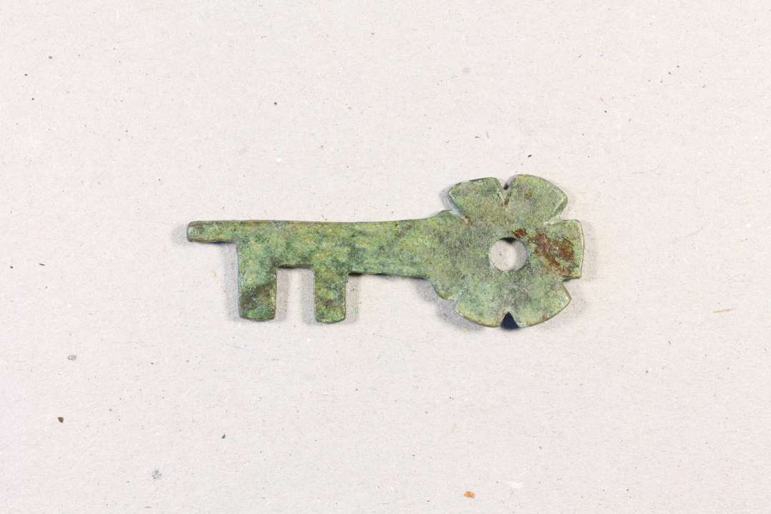 Lille 4,1 cm lang vridnøgle af kobber/bronzeblik, ikke støbt. Hovedet er cirkulært med hul i midten og ligner en blomst. Nøglen er lidt skæv, sikkert af at være blevet vredet rundt i låsen mange gange. Sikkert fra et skrin.