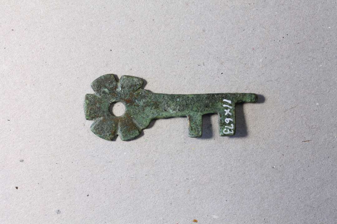 Lille 4,1 cm lang vridnøgle af kobber/bronzeblik, ikke støbt. Hovedet er cirkulært med hul i midten og ligner en blomst. Nøglen er lidt skæv, sikkert af at være blevet vredet rundt i låsen mange gange. Sikkert fra et skrin.