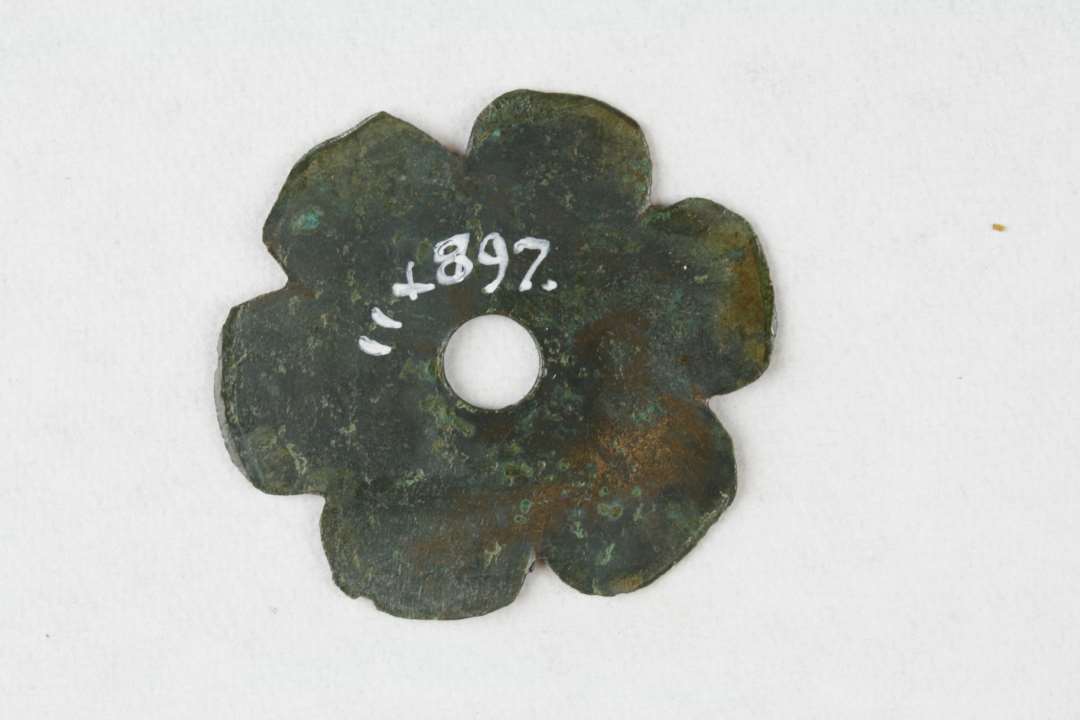 Pyntebeslag af kobber/bronzeblik, formet som en blomst med seks blade og hul i midten, ø 3,5 cm. Kons.