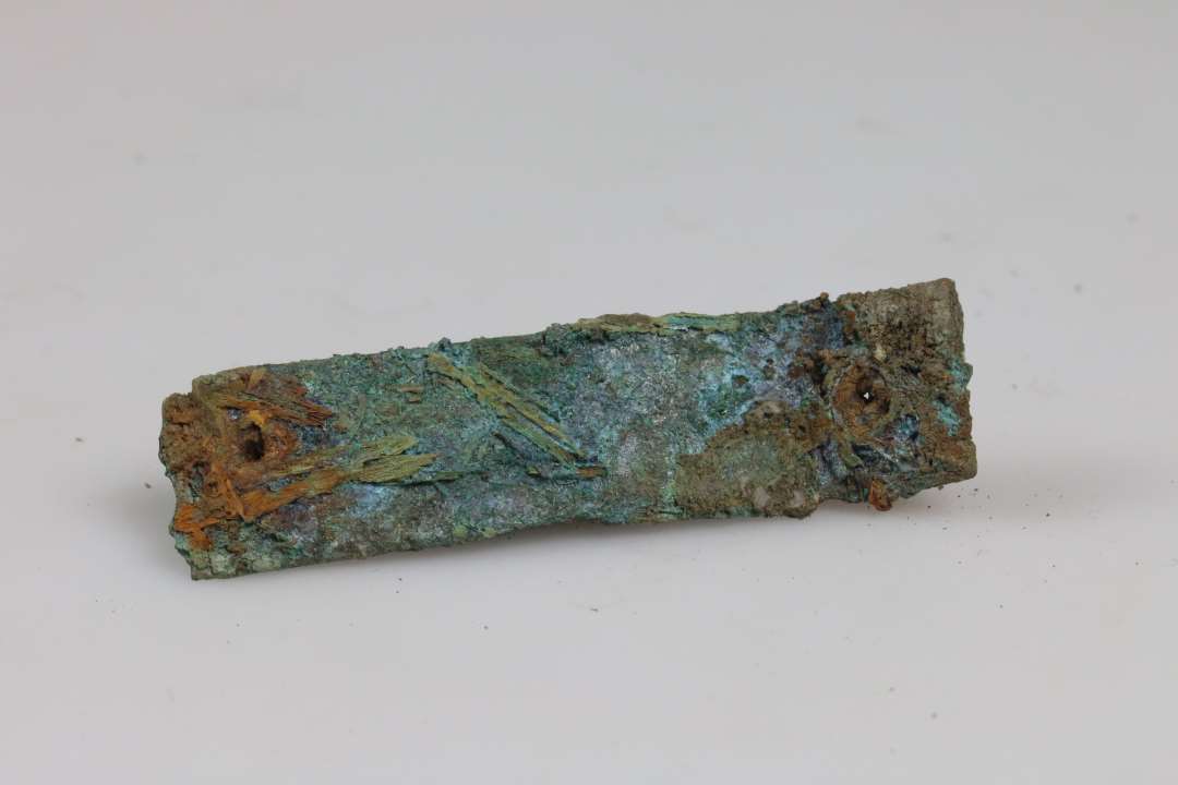 Fragment af beslag? Aflangt, båndformet stykke bronze med en nitte i hver ende. Mål: 7x1,5 cm., Ukonserveret.