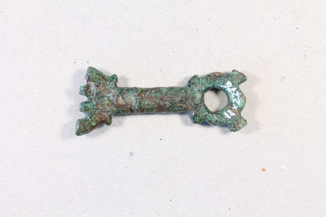 Støbt bronzenøgle, oprindelig med cirkulær fane, der nu er brækket. L. 4,2 cm. Modsat fanen et cirkulært hoved med hul igennem og to små 'ører'