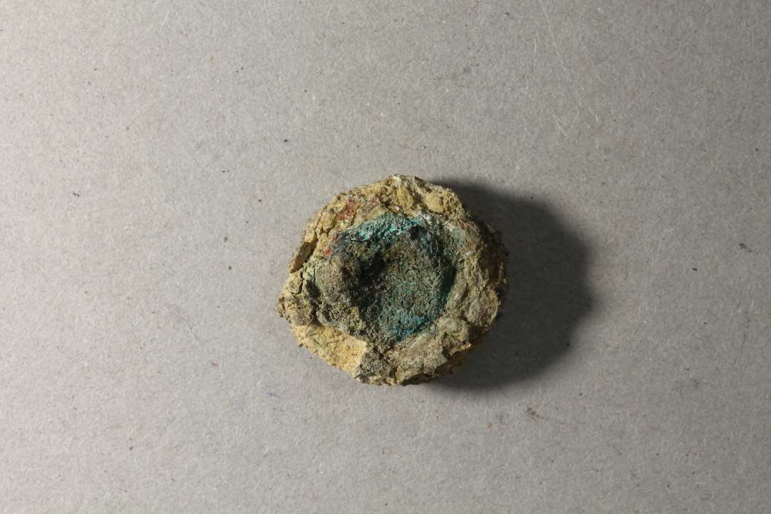 Meget nedbrudt genstand, cirkulær, 2 cm ø, 0,9 cm høj med grøn korrosion i midten. Kan måske være et vægtlod, men det er usikkert