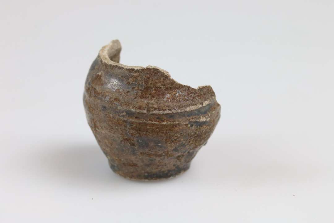 Størstedelen af et miniature lerkar/salvekrukke med flad bund og brun glasur. Bunddiameter: 2,2 cm.