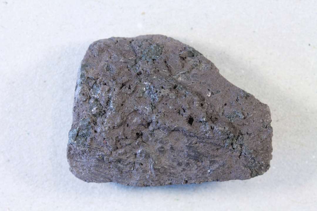 Fragment af noget som muligvis har været en rund, hul ting (diam. måske o. 5 cm). Den øverste kant kan erkendes, men ikke den underste.  Med violet korrosion.