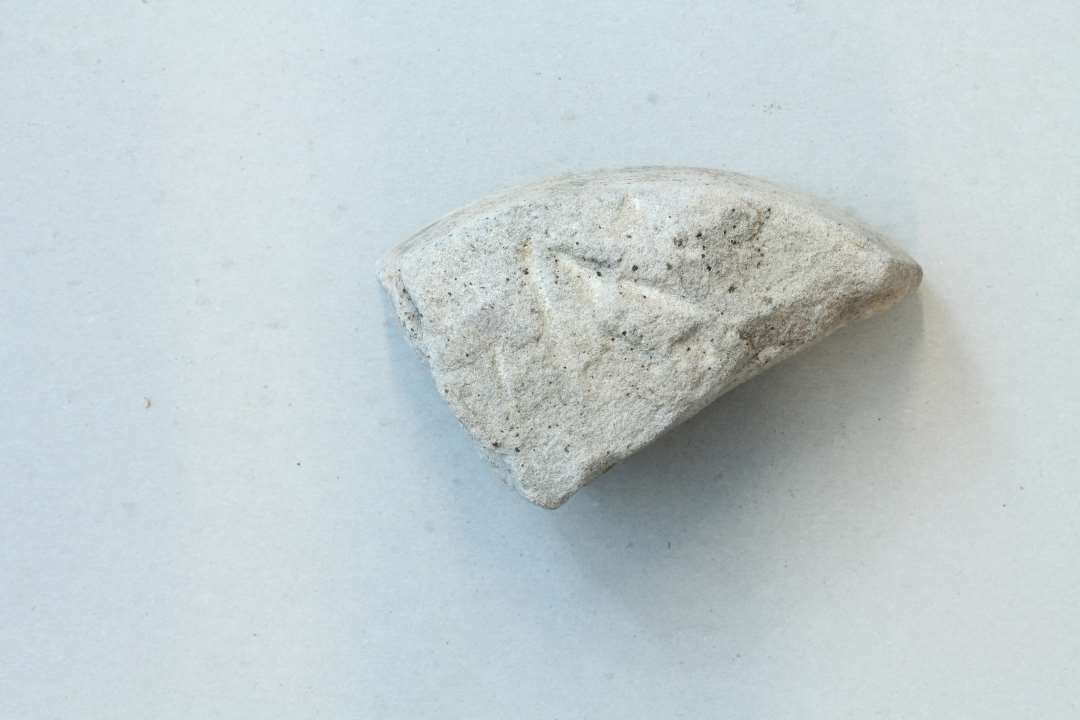 Fragment af cirkulær, roterende slibesten. Der ses rester af den buede slibeflade samt den ene side, der er tilhugget med mejsel. Mål: 9x5,4x5,3 cm.