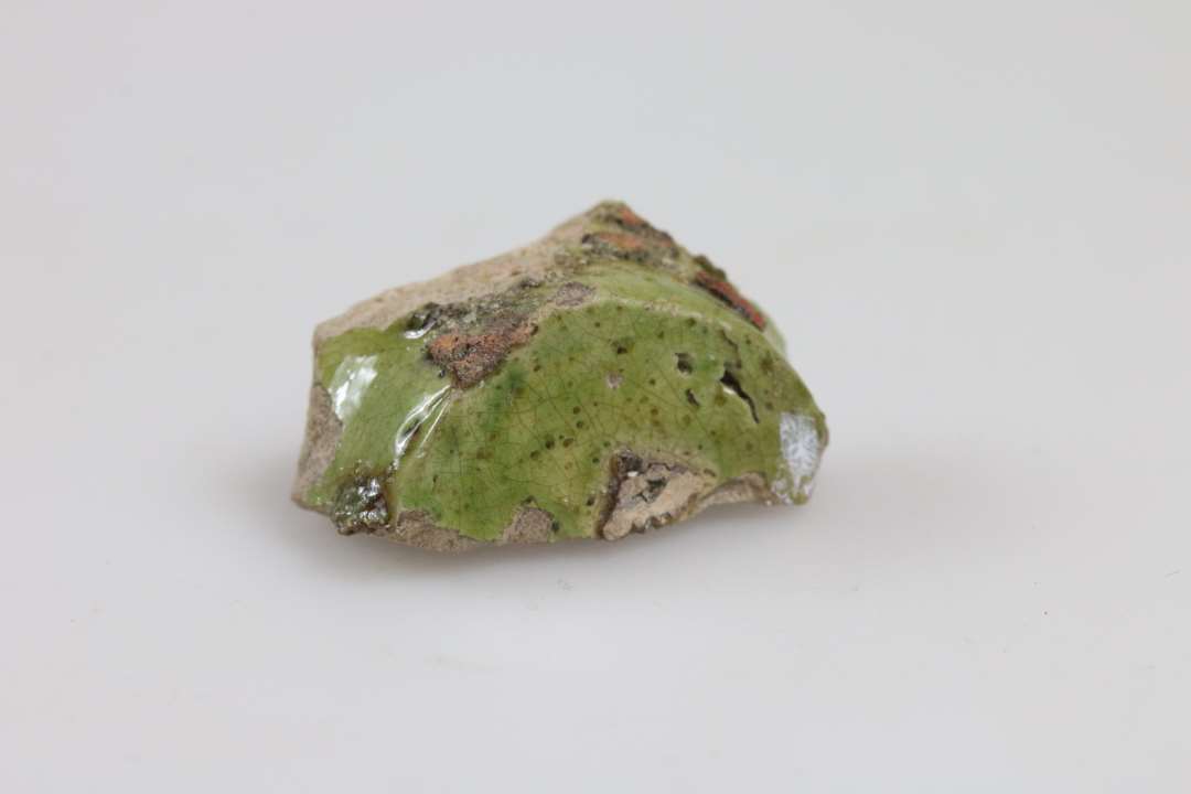 Skår af miniature lerkar/salvekrukke/rangle med flad bund. Hvidt-lyst rødligt gods med udvendig, lysgrøn glasur. Bunddiameter: 2 cm.