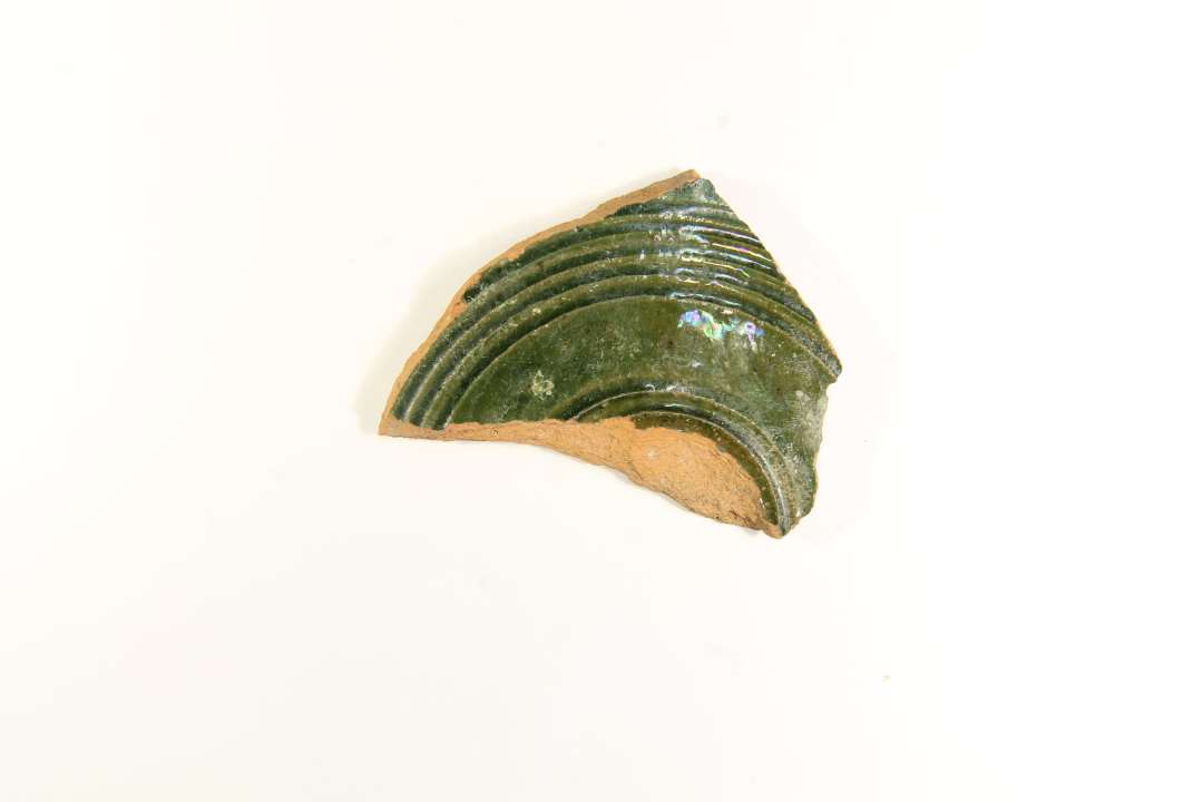 Fragment af grønglaseret, cirkulært lerlåg. Stammer formodentlig fra opbevaringskrukke af finere art, evt. en tobakskrukke Største mål ca. 6 cm.