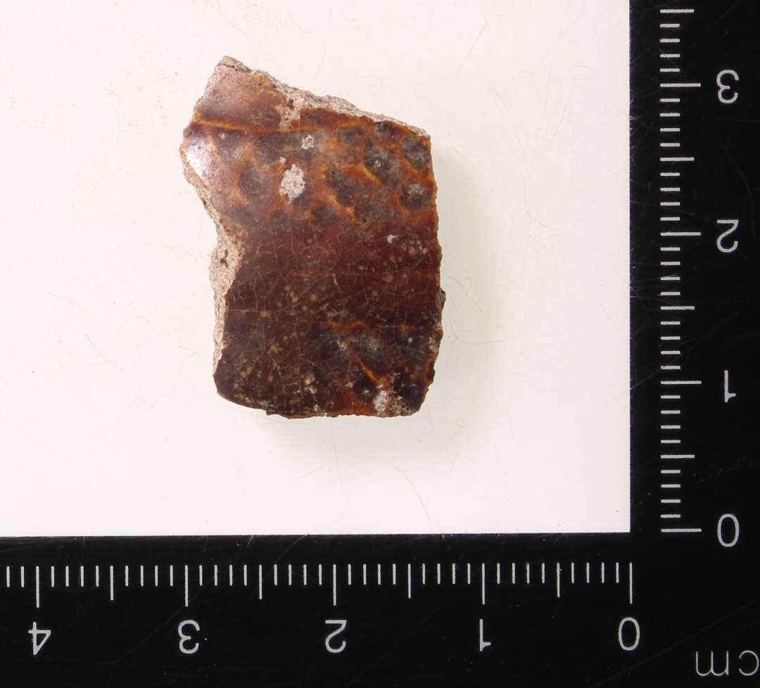 Meget lille, skår med krumning der viser, at det formentlig er fra lille slavekrukke, miniature kar eller lignende. Hvid skærv, rødbrunlig glasur