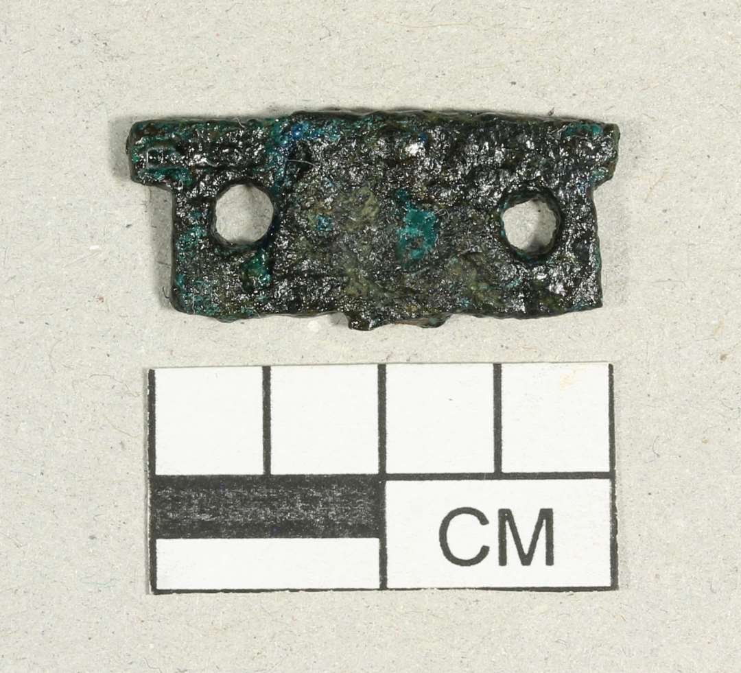 Fragment af beslag. Med to nittehuller. Mål: 2,2x0,9 cm.