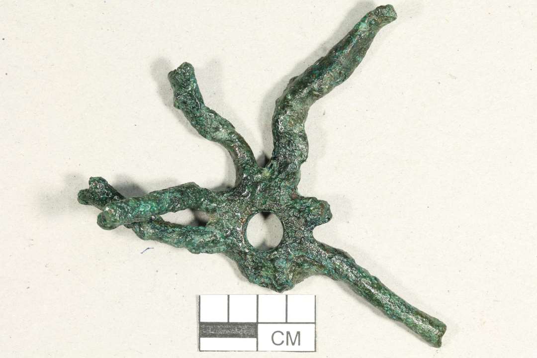 Fragmenteret blæksprutteformet beslag fra skjold. Tidlig middelalder. Noget forvredet. Nittehul ses i en af enderne. Største mål: 7 cm.