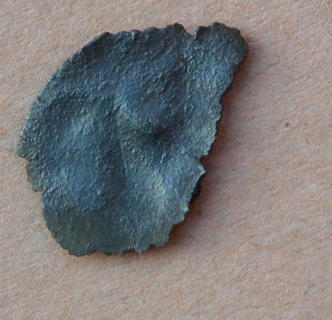 Fragmenteret cirkulært smykke med filigran pelta-figurer omkring centralt punkt. Opr. ca. 27 mm i diameter, største bevarede mål ca. 20 x 16 mm. Bagsiden blank, uden spor efter nåleanordning eller øsken.