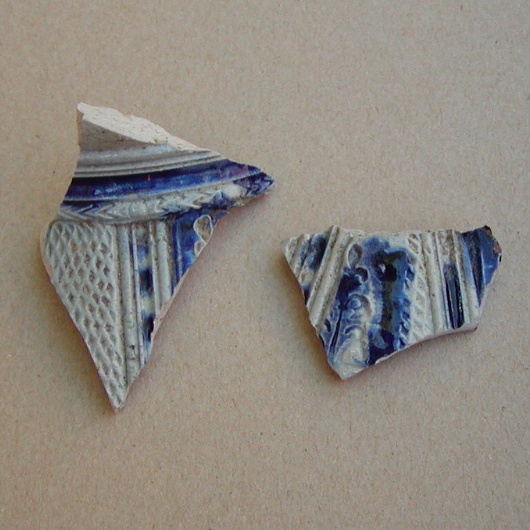 To tinglaserede skår med blå bemaling, blomsterlignende motiv. Det ene et randskår fra et fad, det andet måske fra et krus.