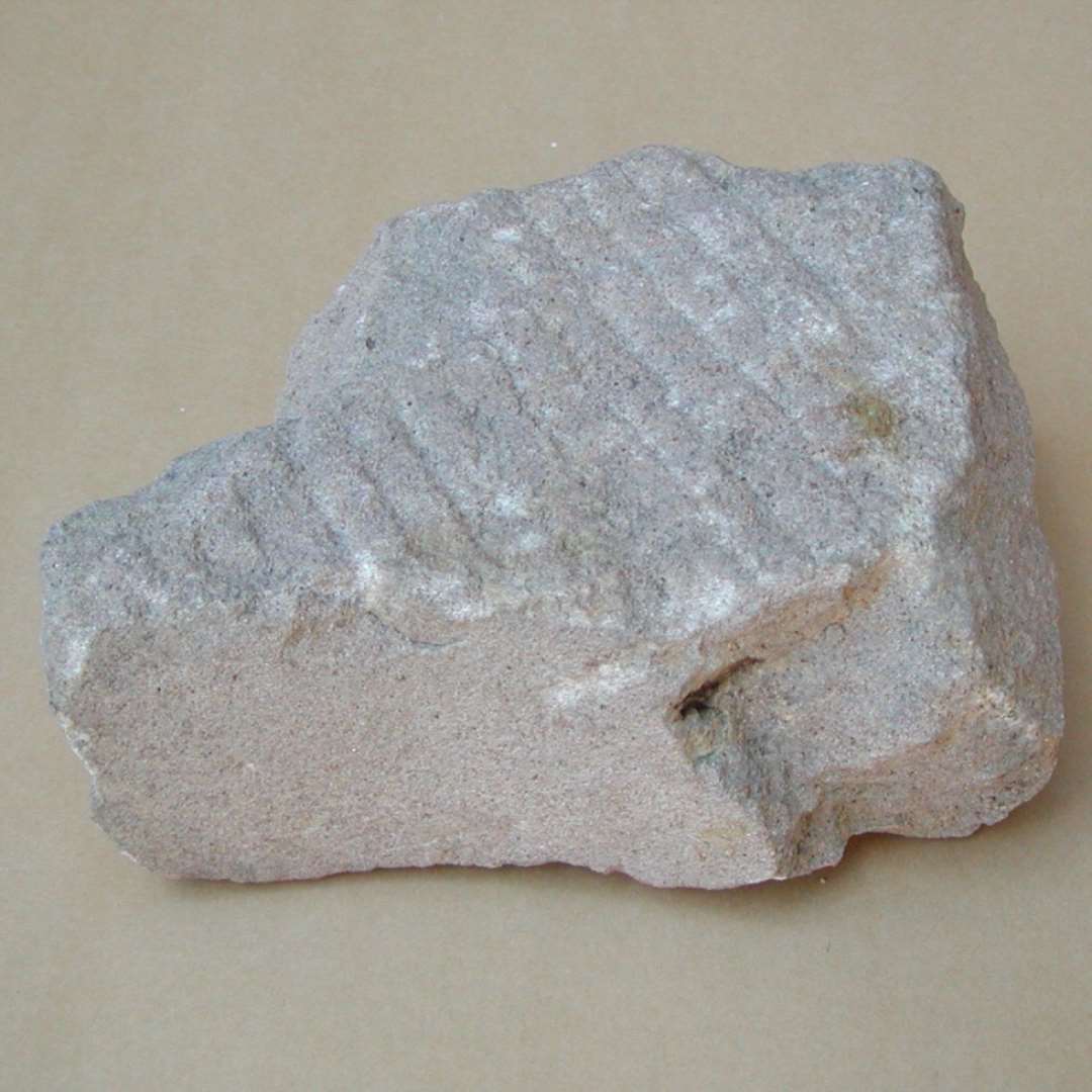 Rødlig sandsten(?) med flere parallelle riller på den ene side. 19x15x8 cm.  Del af sandstenskiste eller låg, måske sekundært anvendt som slibesten på én side.