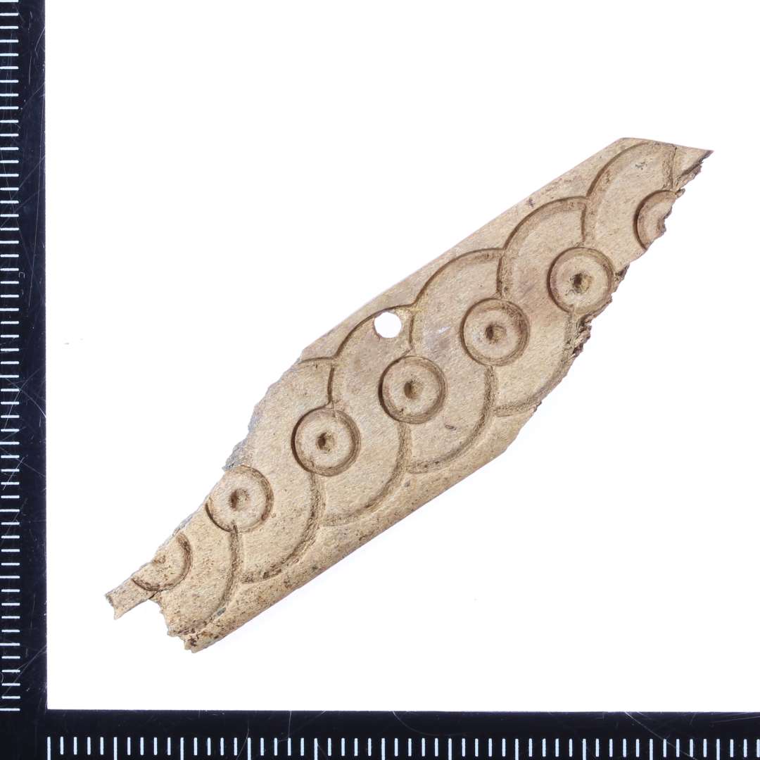 Fragment af pynteplade med 'løbende hund' ornamentik, l. 5,6 cm. Ét monteringshul.