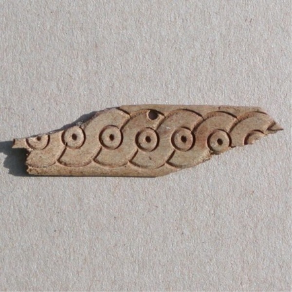 Fragment af pynteplade med 'løbende hund' ornamentik, l. 5,6 cm. Ét monteringshul.
