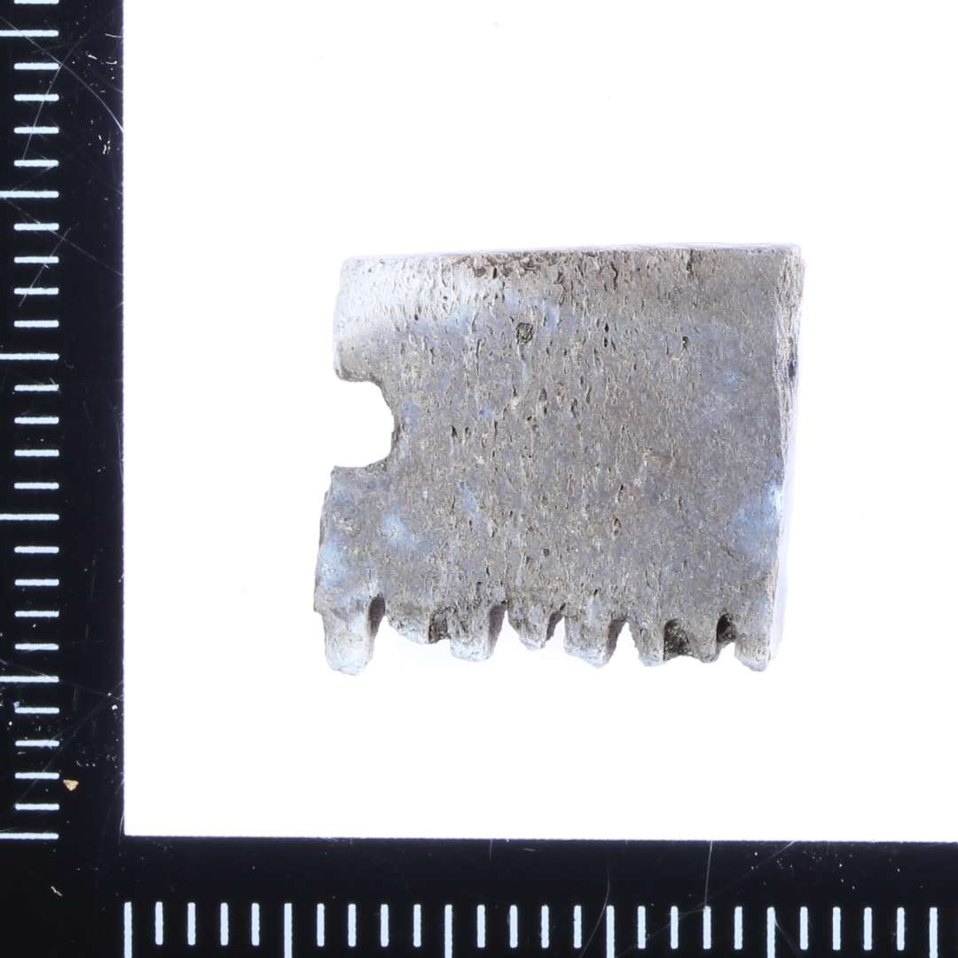 Lille fragment af midterstykke til benkam. Brændt. Rester af afbrækkede tænder på den ene side. Rester af et nittehul i den ene kant.