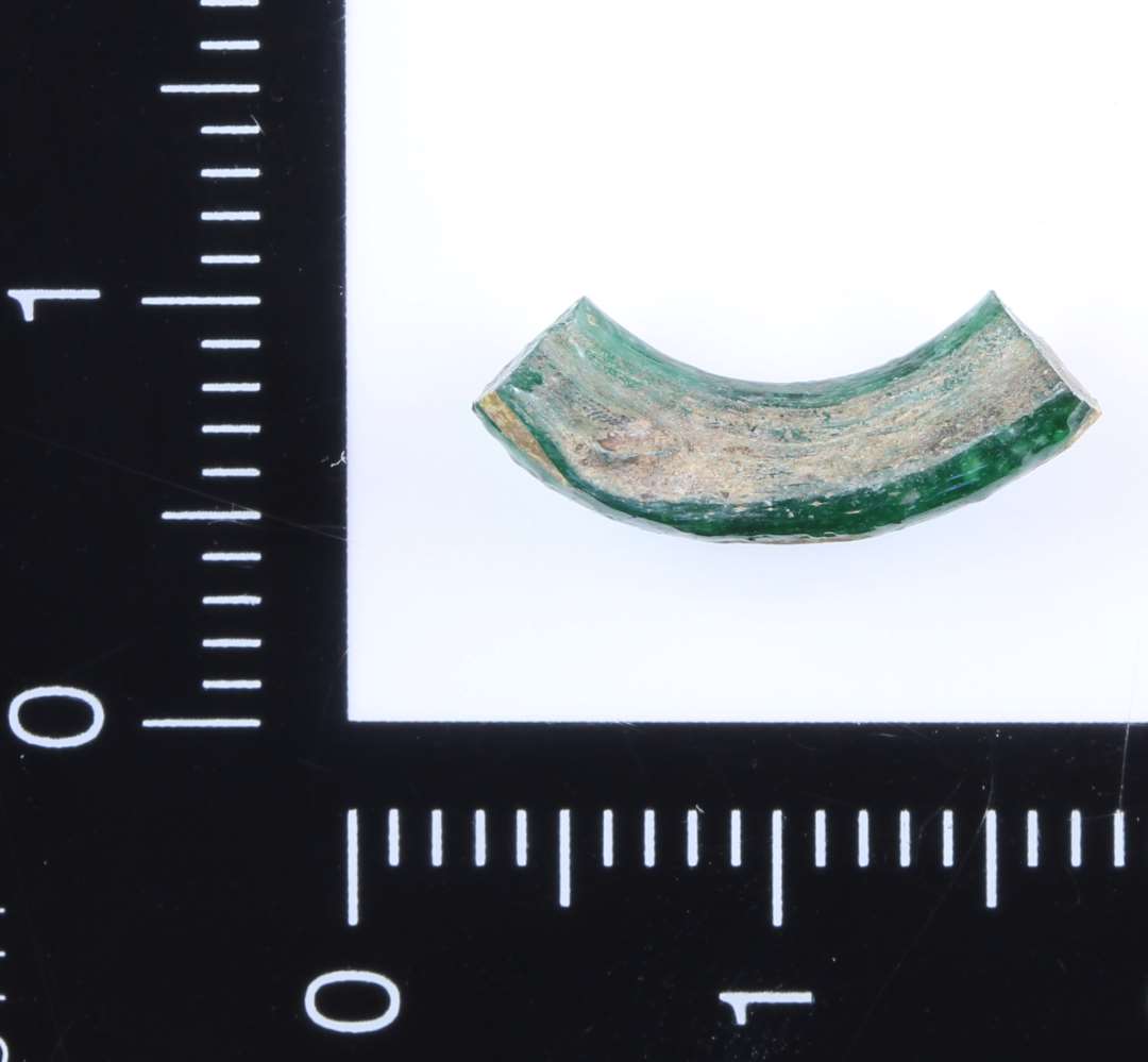 Større fragment af glasring i klart grønt glas. På ydersiden er indlagt bølgede streger af mat hvidgult materiale. Materialet er kun indlagt på overfalden. Formodet diameter på ca. 2 cm.