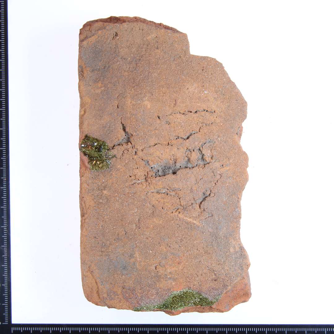 Et  hjørne fragment af en rødbrændt gulvflise med lidt  rester af grøn glasur. Største mål: ca. 13xx7x3,5 cm.