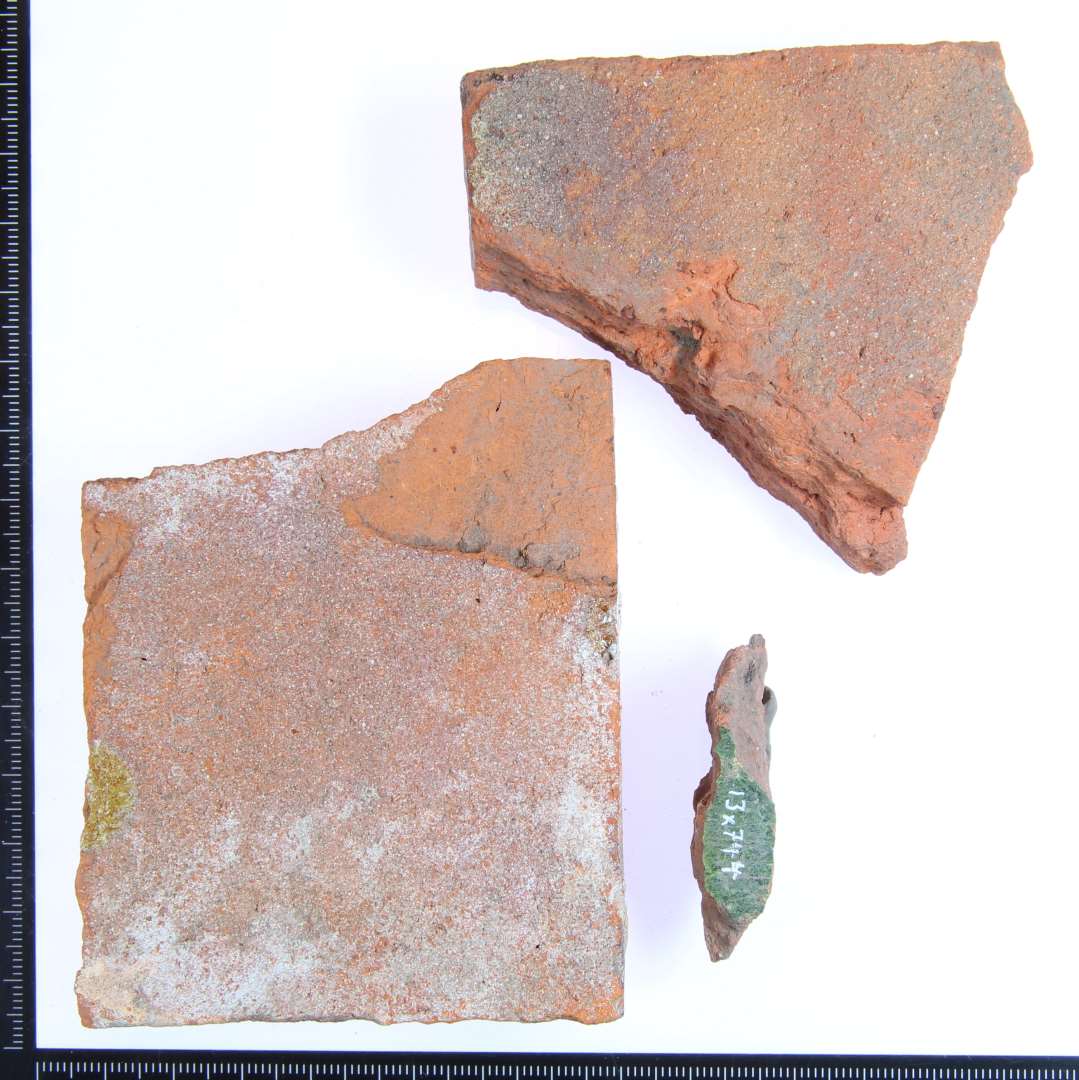 3 fragmenter af fliser. Et meget lille fragment med grøn glasur. Et rødbrændt hjørne fragment med rødlig glasur på en af kantsiderne. Mål:9x7x2 cm. Et rødbrændt hjørne fragment med rødlig glasur på 2 af kantsiderne Mål: 7x6x2 cm.