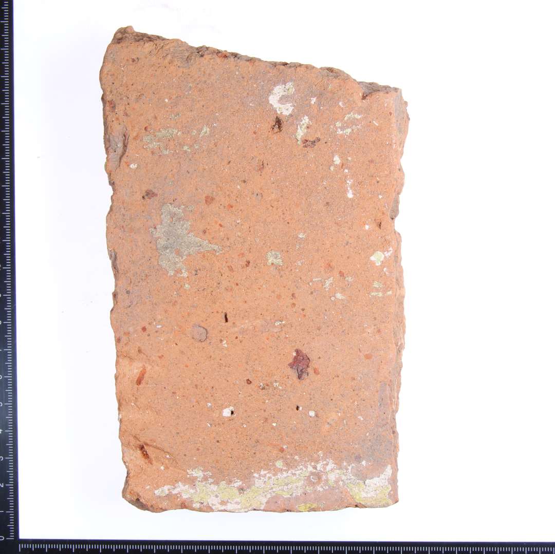 Et hjørne fragment af en gulvflise med lidt gul glasur på oversiden.  Mål: 16x9x2,5 cm.