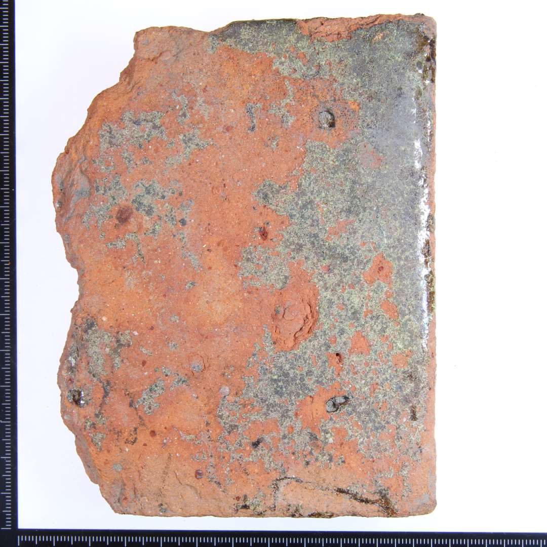 Et rødbrændt hjørme fragment med rester af grøn glasur på overside og alle 3 sidekanter. Største mål: 12x9,5x3 cm.