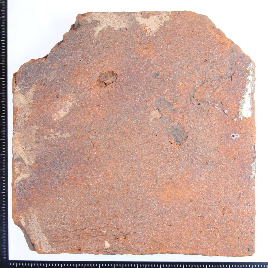 Et rødbrændt kvadratisk gulvflise lettere fragmenteret. Med rester af glasur på 2 af siderne.  Mål: 15x15x2 cm.