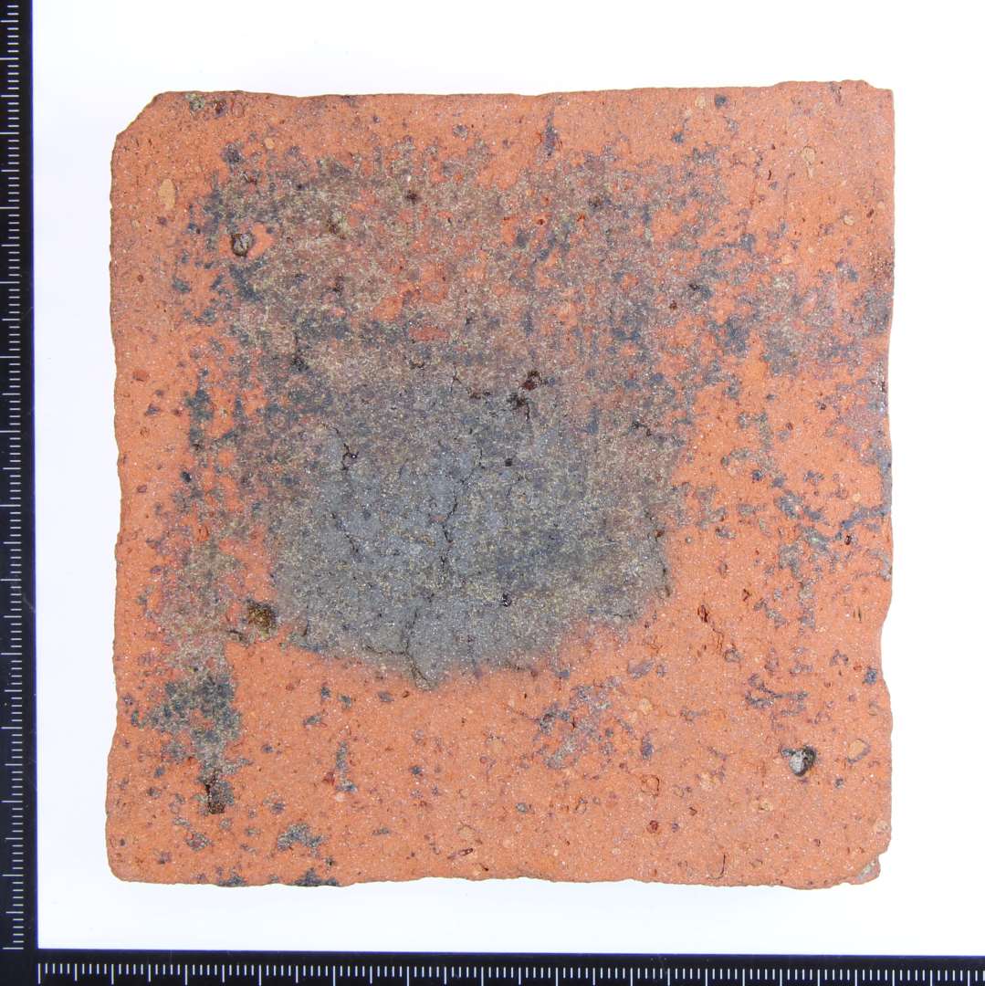 Et rødbrændt kvadratisk gulvflise med rester af mat grøn glasur på oversiden og rødlig glasur på 2 af side kanterne. Mål: 10x10x15 cm. 