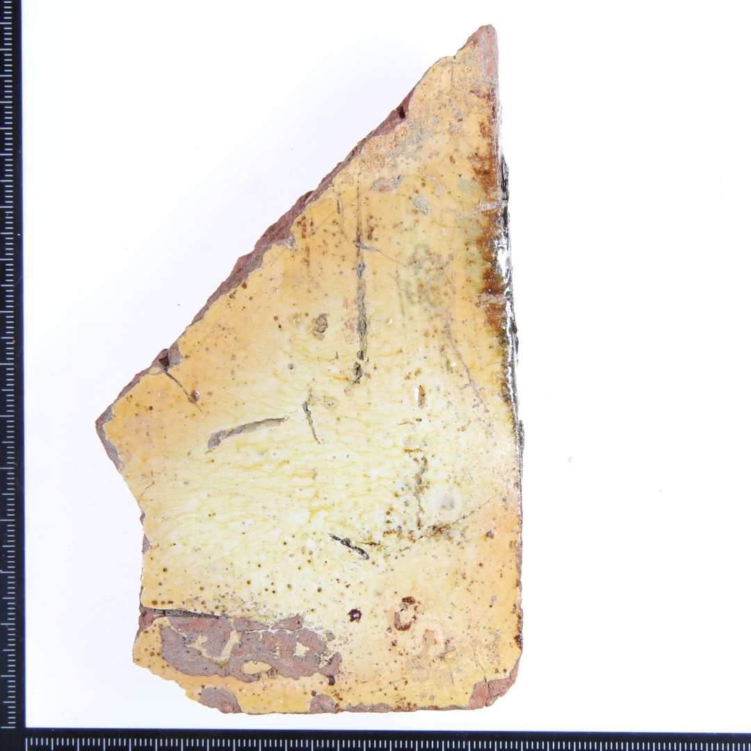 Et rødbrændt hjørne fragment af en gulvflise med gul glasur på oversiden, og rester af rødlig glasur på beggge sidekanter. Mål: 11,5x7x3 cm.