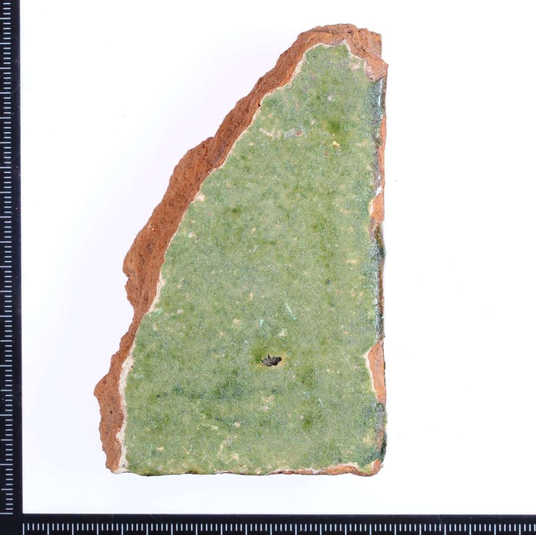 Et rødbrændt hjørne fragment af en gulvflise med mat grøn glasur på oversiden. På de 2 sidekanter rester af grøn/brun/gullig glasur. Mål: 8,5x5x2 cm.  