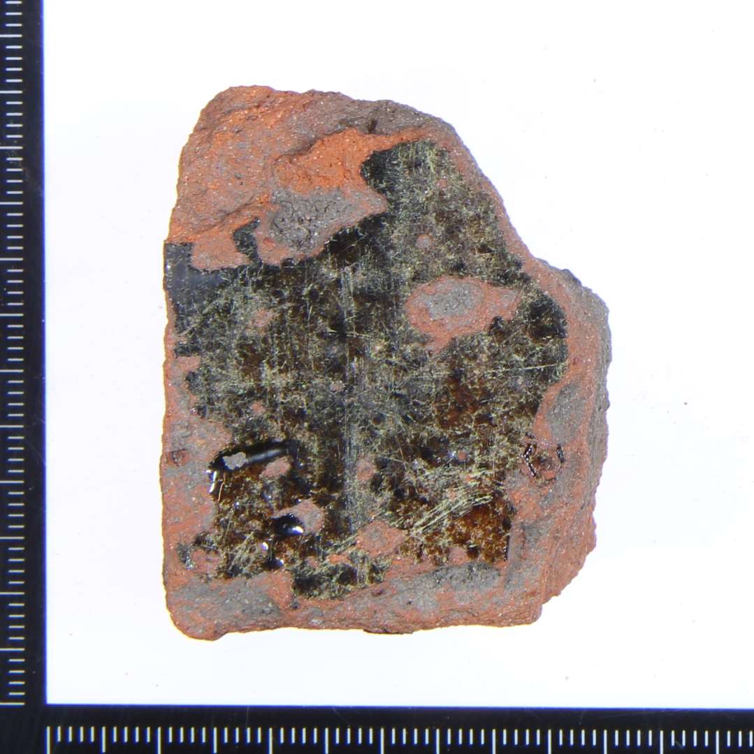 Et lille rødbrændt hjørne fragment af en gulvflise med rester af grønlig glasur på overside og de 2 sidekanter. Mål: 4,5x3x2 cm.