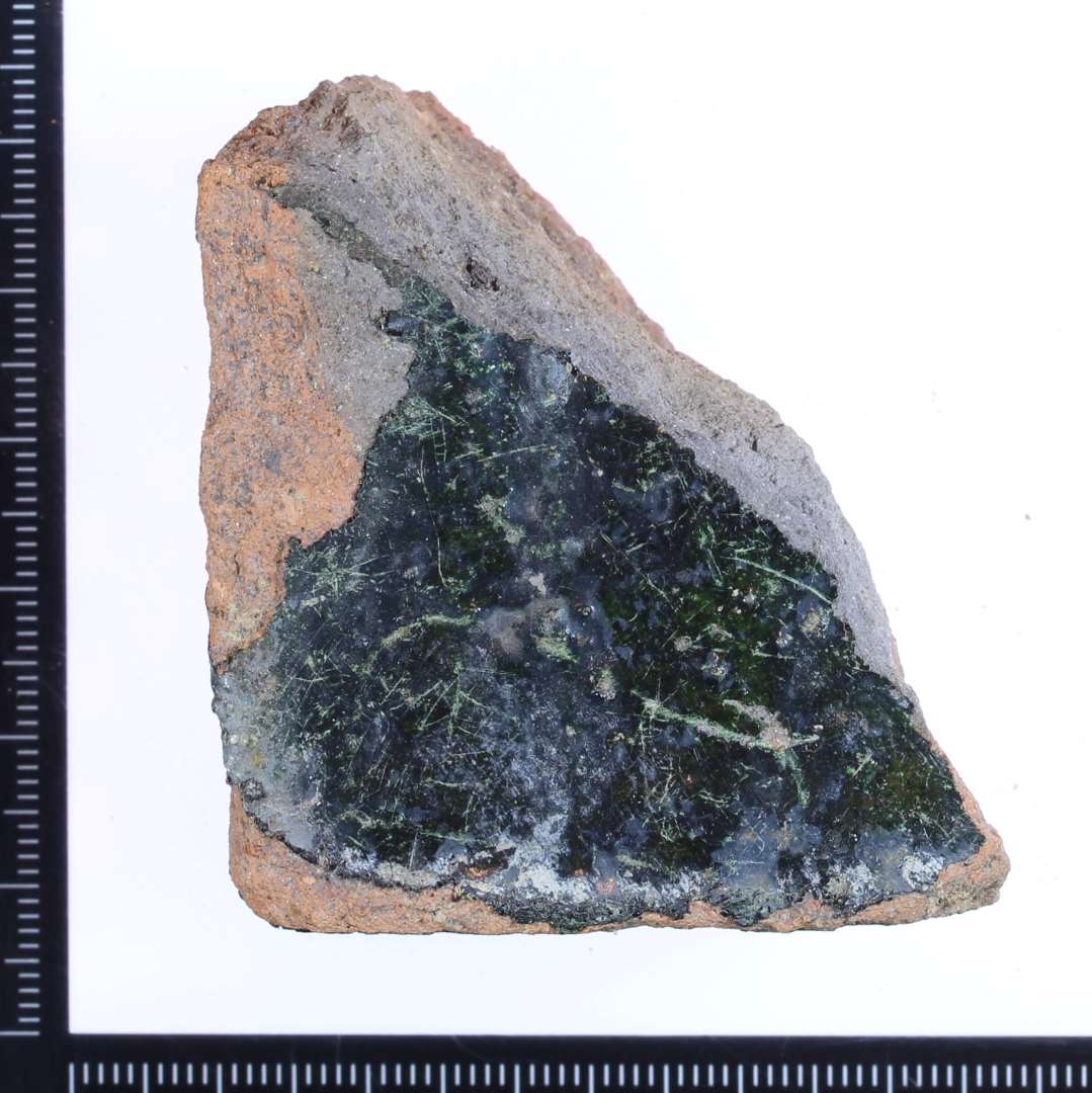 Et rødbrændt hjørne fragment af en gulvflise med rester af grøn glasur på oversiden og begge sidekanter. Mål: 5x5x3 cm.