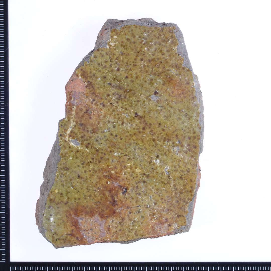 Et rød/grålig brændt fragment af gulvflise?. med brun/grøn glasur på oversiden. Mål: 8x6x1,5 cm.