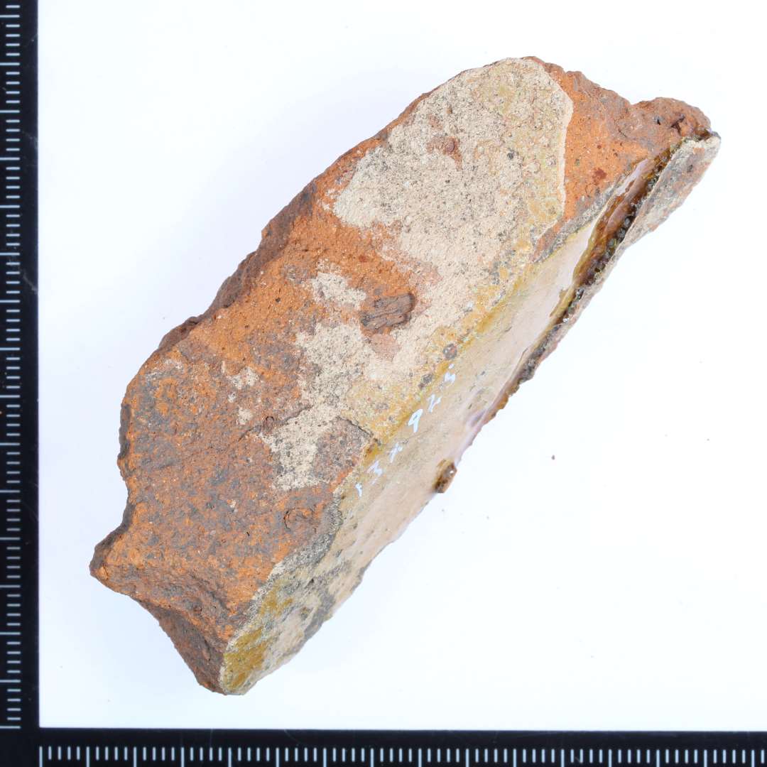 Et lille rødbrændt fragment af en gulvflise med noget af den en kantside bevaret. Rød/gullig glasur på oversiden, og på kantsiden hvidlig rester?. Mål:7x2,5x2,5