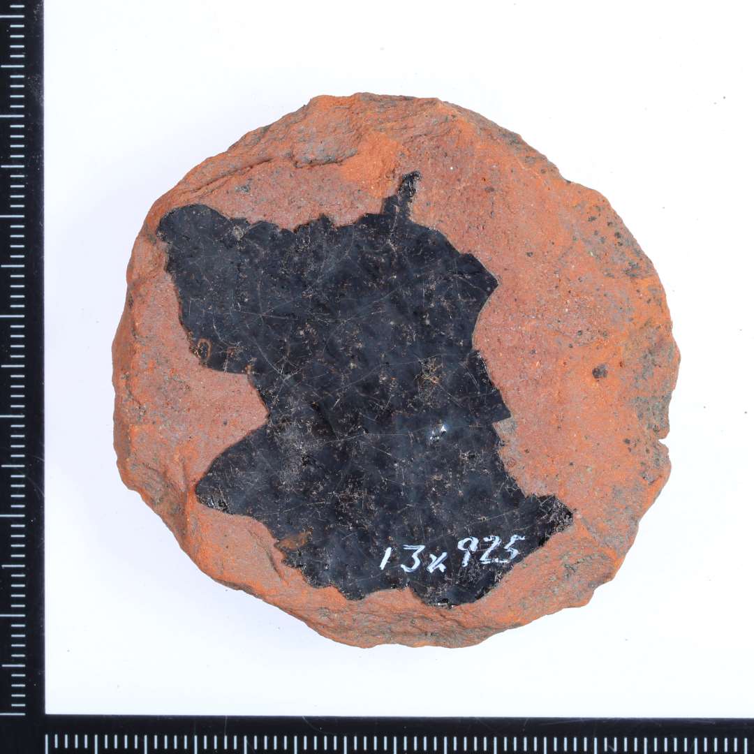 Et rødbrændt fragment af en gulvflise? med rester af mørkgrøn glasur på oversiden. Mål i diameter: 5 cm.
