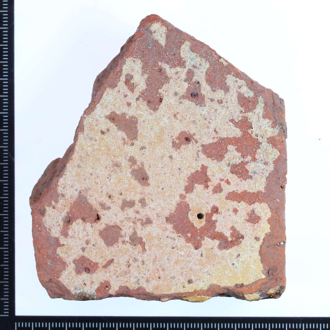 Et rødbrændt hjørne fragment af en gulvflise.På oversiden rester af gullig/hvidlig glasur?, på den ene kantside brunlig glasur og på den anden kantside rester af grøn mat glasur. Mål:8x7x2,5 cm.